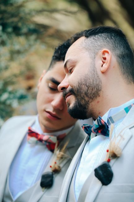 The image depicts two men in close proximity with their foreheads gently touching, both dressed in formal attire suitable for a wedding or special event. The man on the right has a neatly groomed beard, short dark hair, and wears a light-colored suit with a white shirt and a multi-colored bow tie. The man on the left has a similar hairstyle, also with a suit jacket, white shirt, and a bow tie, and appears to be slightly younger. Both men have boutonnières pinned to their lapels, indicating a celebratory occasion. The background features blurred natural greenery, suggesting an outdoor setting, possibly in a park or garden, with soft, diffuse lighting enhancing the intimacy of the moment. The overall scene radiates tenderness and connection, capturing an emotional moment, with a focus on their facial expressions and attire. This image reflects the celebration of a union, emphasizing personal relationships over the surrounding environment, fitting within themes of private ceremonies or same-sex wedding customs. This detailed visual description supports accessibility and aligns with content related to personal events and wedding services.