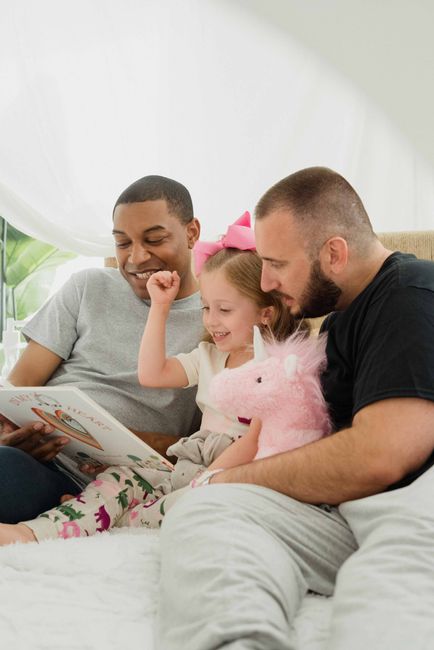 A family sitting closely together on a sofa indoors, with two adults and a young girl, sharing a moment of joy while reading a book. The young girl, wearing a pink bow in her hair, is smiling and raising her fist in excitement, holding a plush pink unicorn toy on her lap. The man on the right, with a beard and short hair, is wearing a black t-shirt, and is leaning in towards the girl, engaged in the activity. The woman on the left, with short hair and a grey t-shirt, is smiling warmly as she looks at the book. The background includes a white curtain and some green houseplants, contributing to a bright, comfortable atmosphere. The scene exemplifies a joyful, family-oriented moment, with an emphasis on creating a warm environment, akin to the comforting setting of a home, similar to the surroundings where rubbish removal services like those provided by Waste Clearance Bow could be performed in a domestic context.