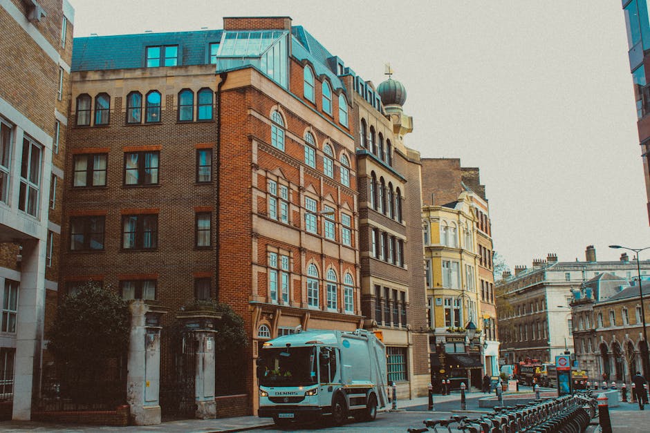 A city street scene featuring a mix of historic and modern buildings, including a prominent multi-storey brick building with arched windows and decorative stonework, set against a pale sky. In the foreground, a small waste collection truck with a blue cab and white body is parked near the curb, indicating recent rubbish collection activity. Surrounding structures include a beige and light blue building with bay windows, a white stone building with classical architectural details, and a modern glass building on the right. The street is lined with black bollards and bicycle racks, and a few pedestrians can be seen in the distance. The vehicles and infrastructure suggest an urban environment where private waste collection and rubbish removal services are utilized, supporting the context of alternative waste handling solutions in central London.