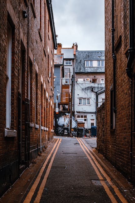 A narrow urban alleyway flanked by red brick buildings on both sides, with the paving marked by double yellow lines along the edges. The alley leads to a back area at the rear of the buildings, where a black waste collection bin is visible among other waste containers. The background features a grey concrete building with multiple windows and a weathered appearance, with some piping and small external structures attached. The scene appears damp, with overcast sky above, suggesting recent rain, and subtle natural lighting casting soft shadows. This setting may relate to private waste handling or alternative rubbish removal options, in line with independent collection practices often used for local property clearances, such as those carried out by waste management services like Waste Clearance Bow.