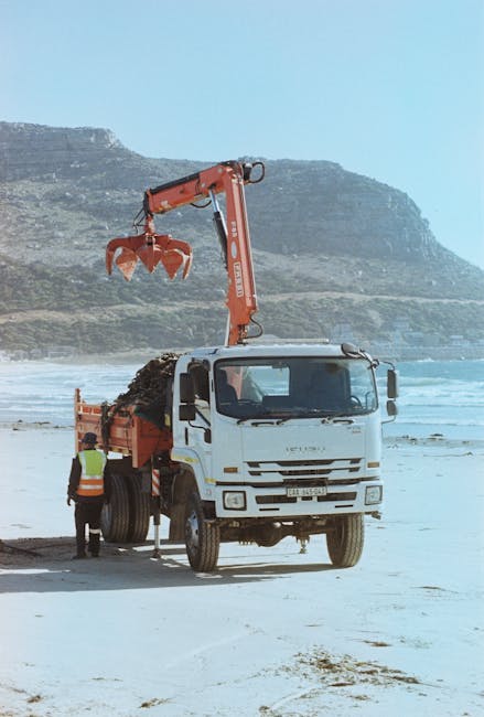 A white waste collection truck equipped with a large hydraulic arm and orange grapple attachment, positioned on a sandy beach with the ocean and a rugged, grassy cliff in the background. The truck's cab is clean with a large front windshield and side mirrors, and the body is fitted with a large open bed loaded with dark, mixed waste material. A worker dressed in high-visibility clothing is standing near the side of the truck, overseeing the loading process. The scene is outdoors during daylight, with natural light illuminating the truck and surroundings, reflecting the typical environment for private waste handling or alternative rubbish removal services, such as on-site clearance away from public refuse collection points. The background shows calm ocean waters and a film of sand along the shoreline, indicating a coastal location where independent waste clearance activities might be taking place without reliance on local authority collection. Waste Clearance Bow would facilitate such on-site waste removal, managing rubbish in varied environments like beaches or private properties.